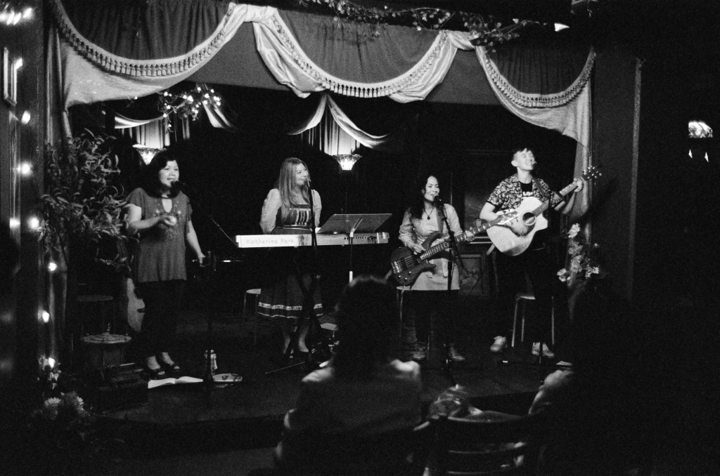 Left to right on a lit stage with Victorian tasseled curtains, @francesanchetasongwriter shakes an egg shaker and sings into a mic, @katherinepark sings into the mic at the keyboard looking up, @lisagraciano sings into her mic and plays her electric bass, @mjoyourselfmusic sings into her mic with eyes closed and plays acoustic guitar at our first gig together, with two women in the front row backs to the camera, “AAPI Songwriters in the round” at @thelostchurchsf in San Francisco , California on 7 July 2023 , photographed by @douglasdespres .